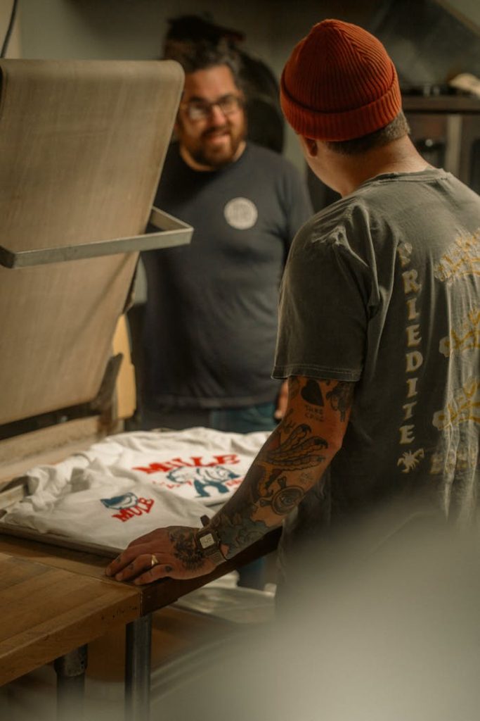 The Art of Drawing Readers In: Your attractive post title goes here Two men engaged in screen printing a t-shirt in a cozy workshop setting.