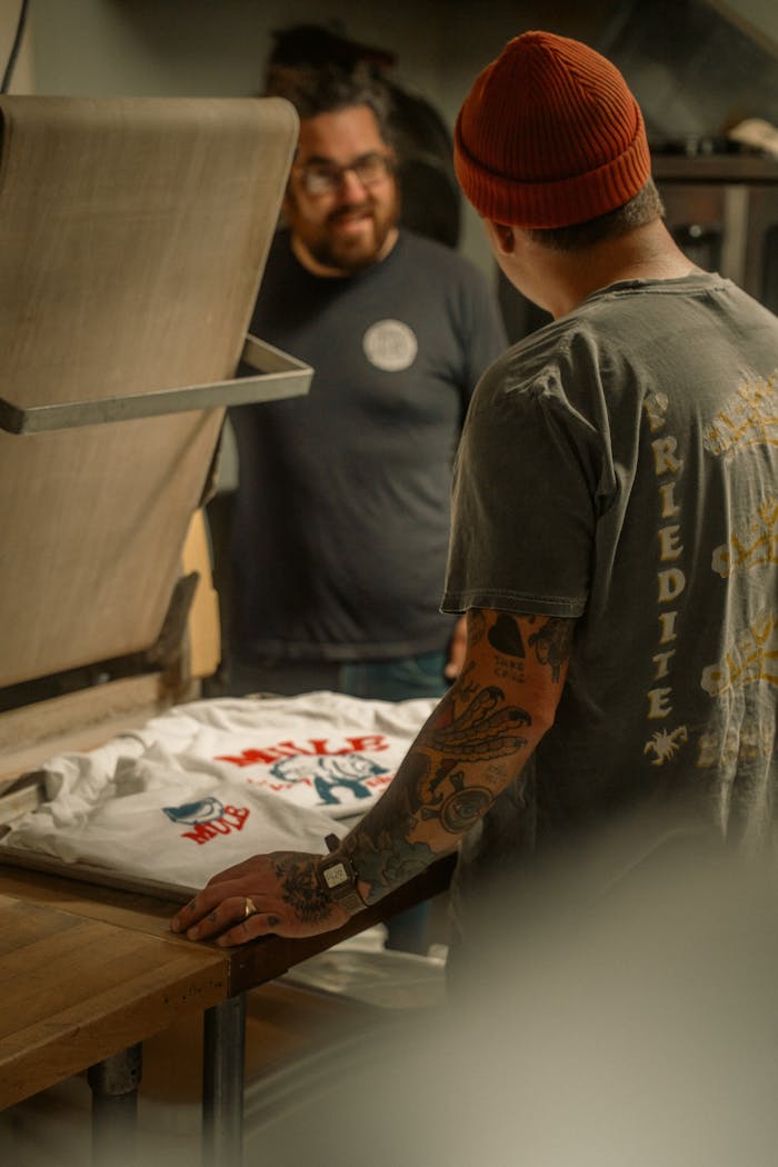 Two men engaged in screen printing a t-shirt in a cozy workshop setting.
